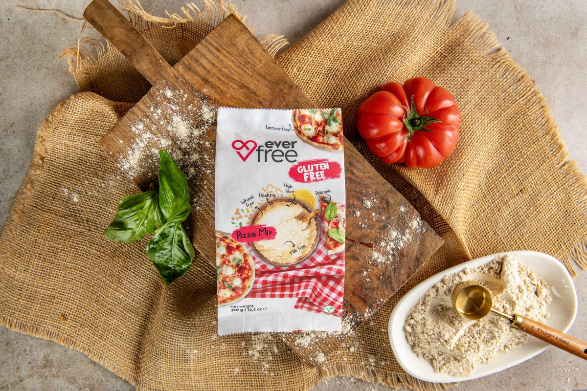 EverFree Gluten-Free Pizza Mix bag on a wooden board with fresh basil, a tomato and a bowl of the dry mix